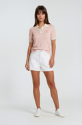 Woman wearing a light pink birdies and bubbles knit polo shirt and white shorts on a white background