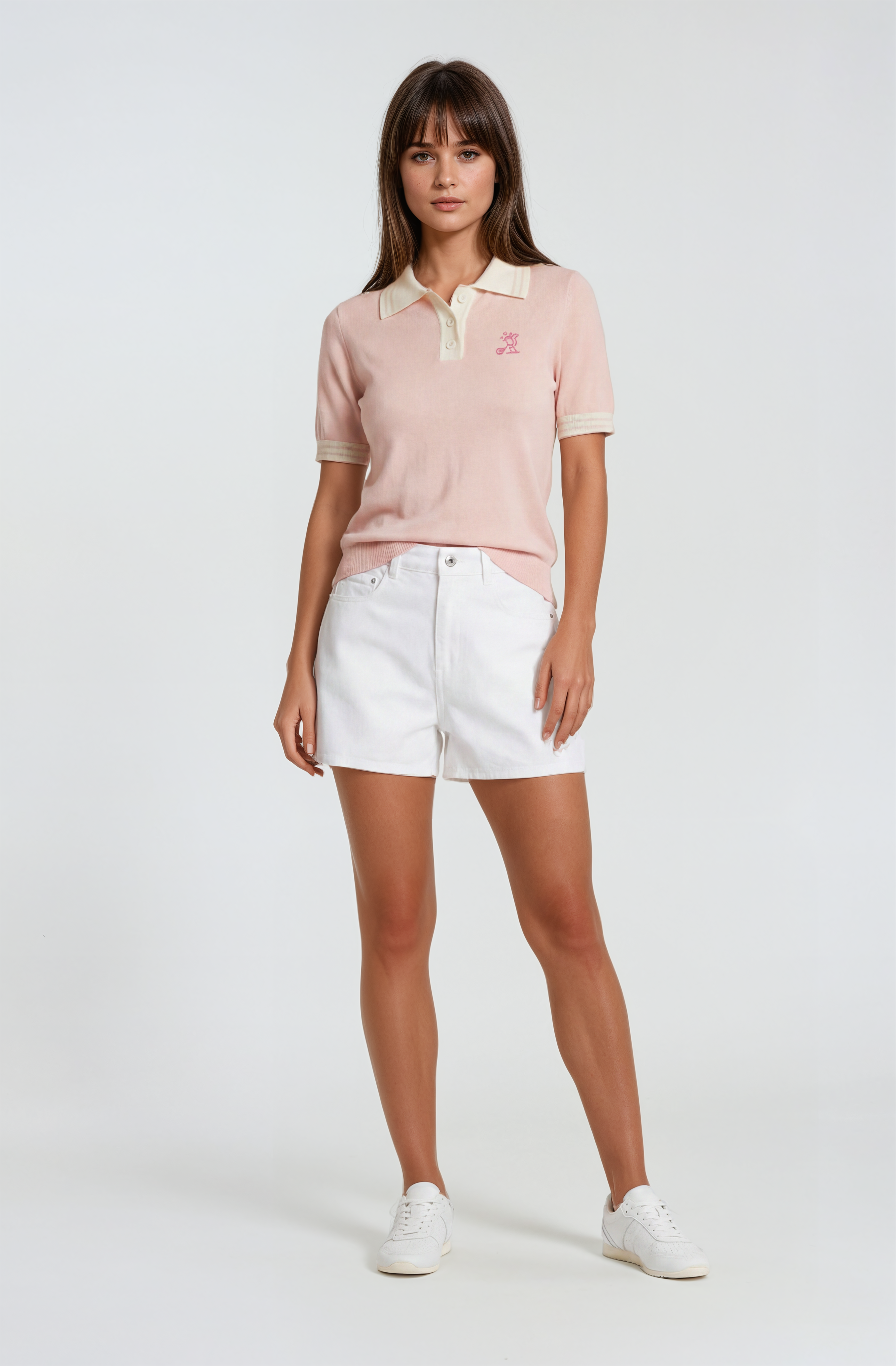 Woman wearing a light pink birdies and bubbles knit polo shirt and white shorts on a white background